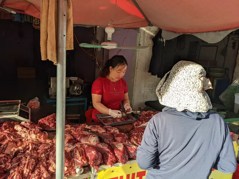 Butcher selling raw meat at an outdoor market stall, serving a customer wearing a hat.