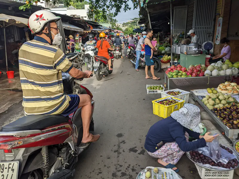 Busy street market with fruit vendors, shoppers, and motorbikes passing by.