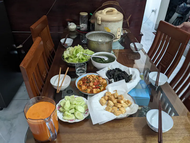 Vietnamese family meal with fried tofu, stuffed betel leaves, stir-fried vegetables, lettuce, soup, and carrot juice on a dining table.