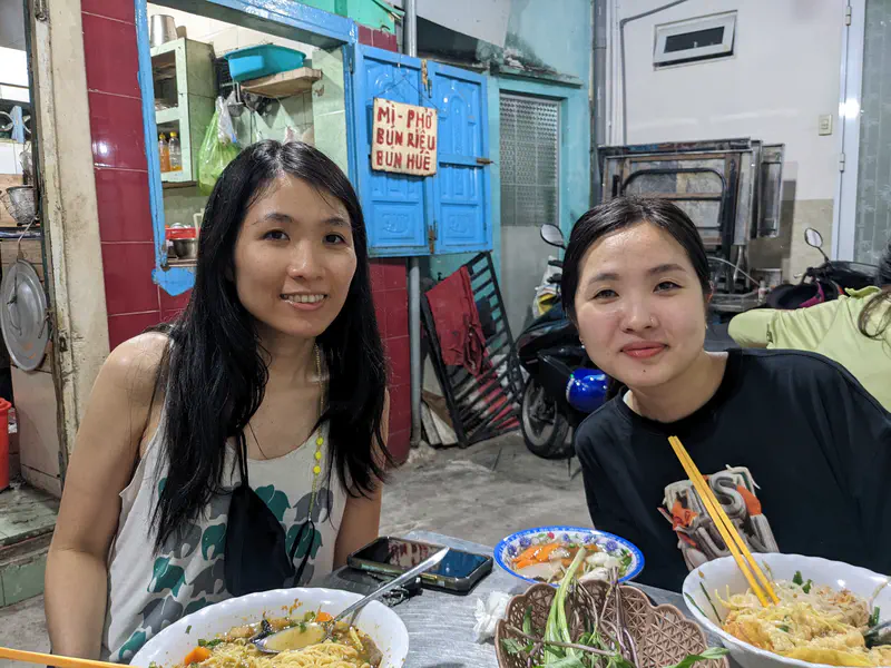 Two women sitting at a casual eatery table with bowls of noodle soup and vegetables.