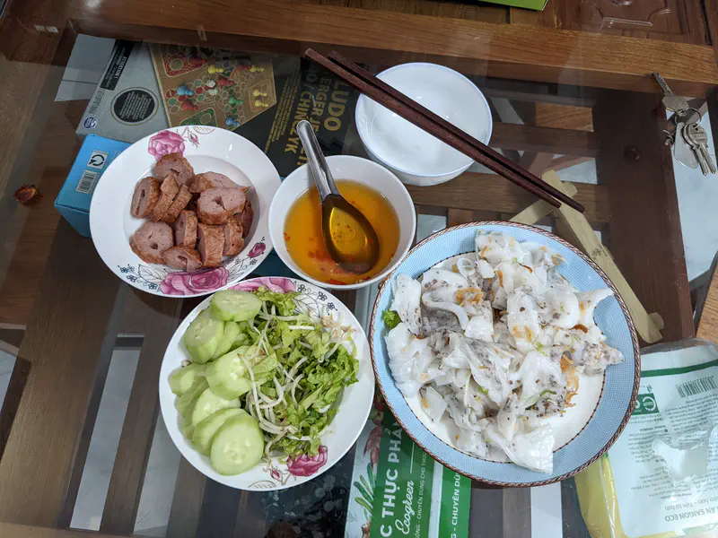 Vietnamese meal with sliced sausage, dipping sauce, cucumber salad, and a plate of steamed rice rolls with filling.
