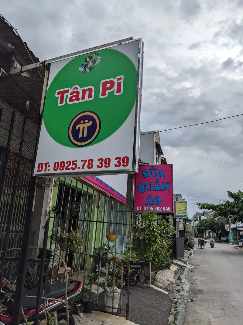 Street with signs reading 'Tân Pi' and 'Sửa Quần Áo' beside small shops.
