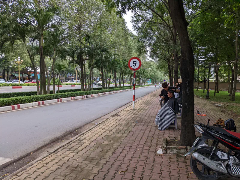 Outdoor barber cutting a man’s hair on a sidewalk beside a tree-lined street.