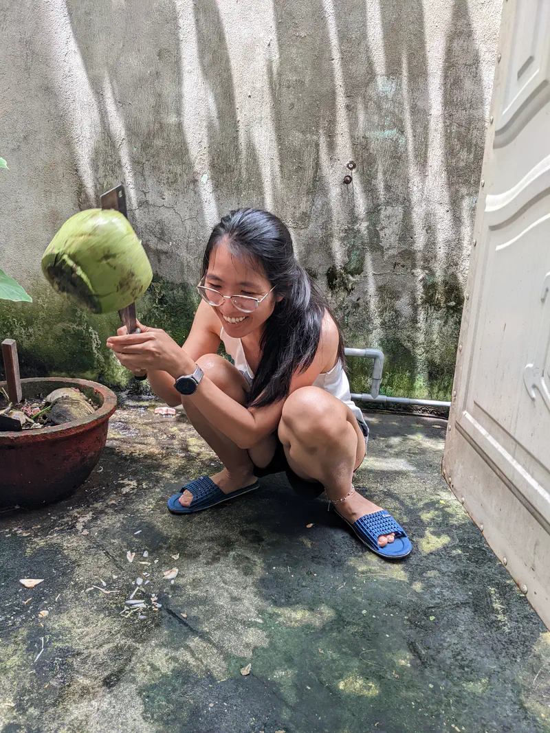 Smiling woman squatting outdoors, holding a green coconut against a blade to open it.