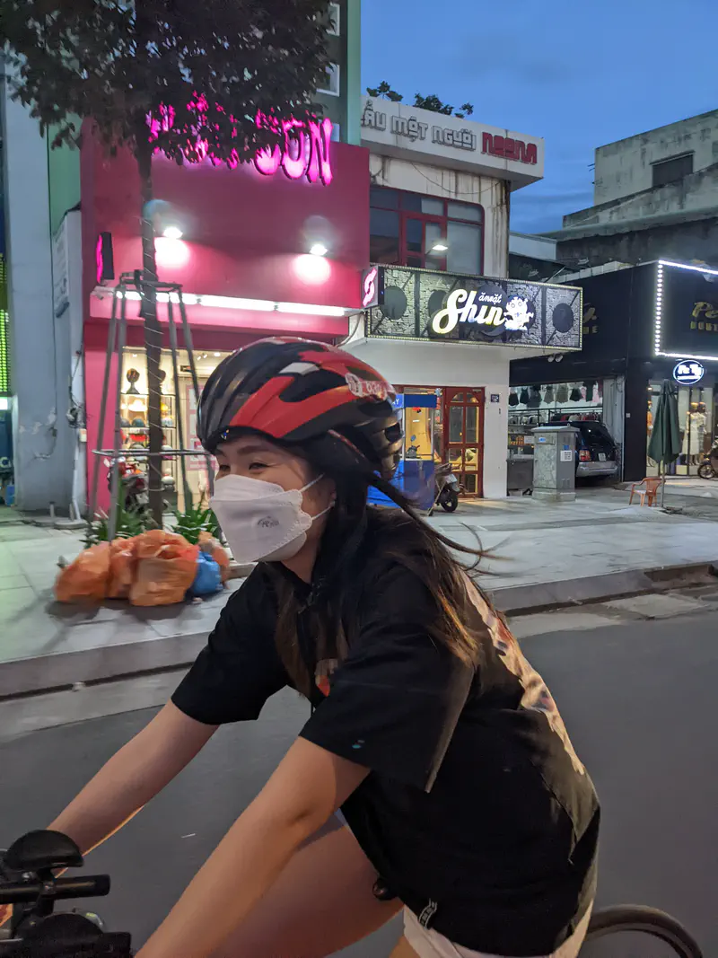 Young woman wearing a mask and helmet riding a bicycle on a city street at dusk.