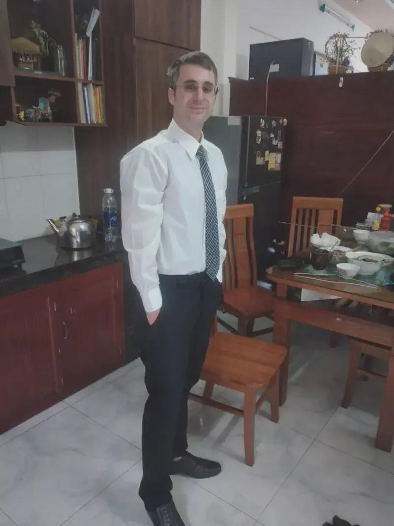Man in formal attire with white shirt and tie standing in a kitchen near a dining table.
