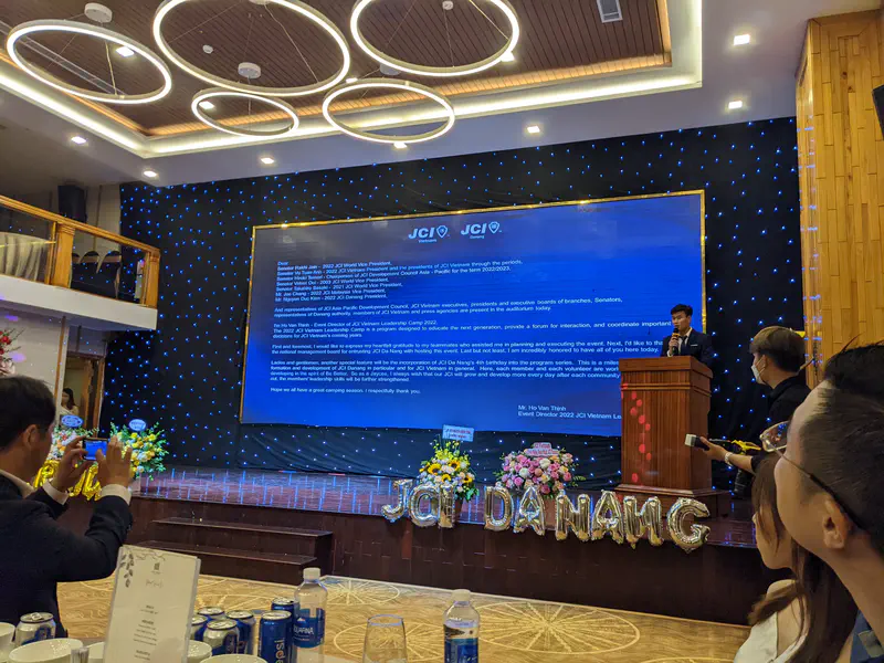Speaker presenting at the 2022 JCI Vietnam event in Da Nang with a large screen displaying text and audience members watching.
