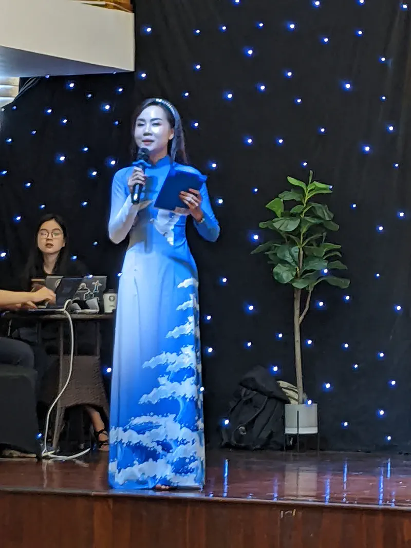 Woman in a blue áo dài speaking on stage with a microphone and holding notes, with a starry backdrop behind her.