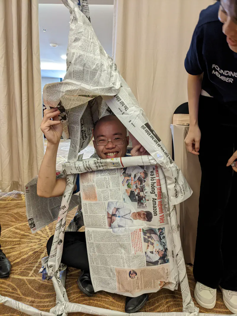 Smiling man crouching under a structure made of newspapers during an activity.