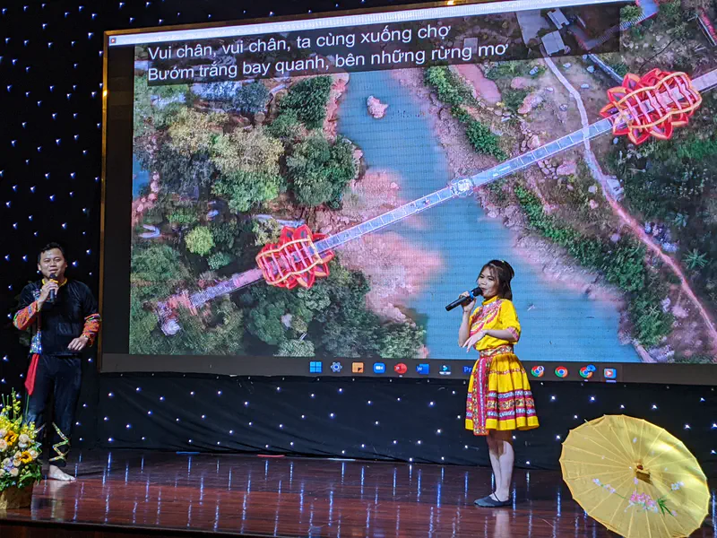 Two performers in traditional costumes singing on stage with a large screen showing an aerial view of a bridge and Vietnamese lyrics.