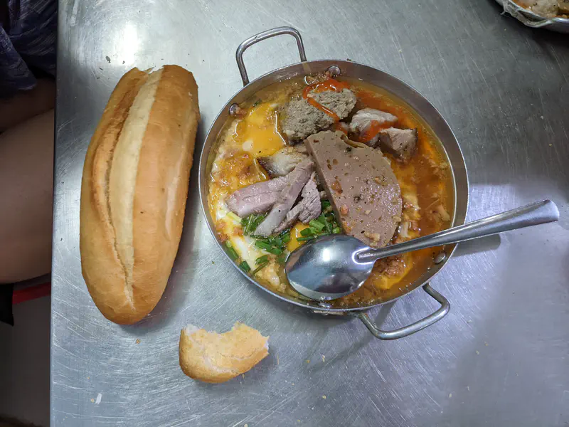 Pan of Vietnamese dish with meat, egg, and sauce served alongside a baguette.