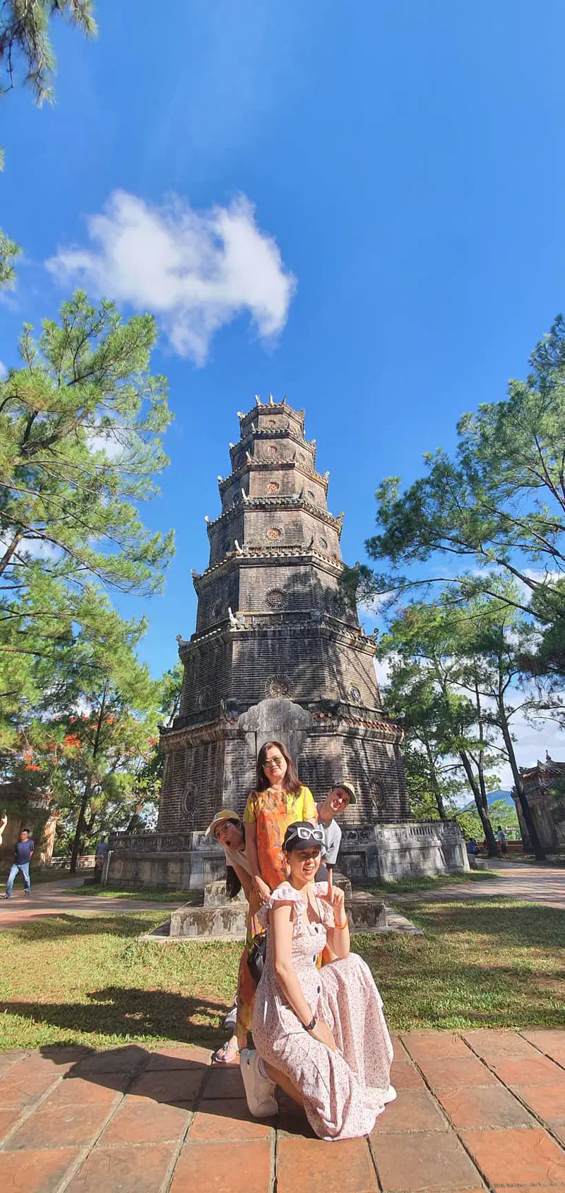 Group of people posing in front of the historic Thien Mu Pagoda in Hue, Vietnam, with a clear blue sky and tall trees around.