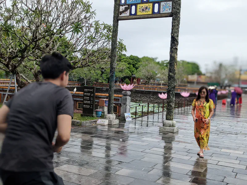 Man taking a photo of a woman walking on wet stone pavement near a decorated gate in Hue.