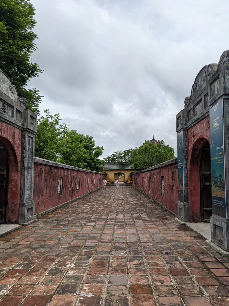 Stone walkway leading through a red-walled gate toward another historic building in Hue.