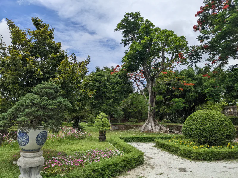 Lush garden with bonsai trees, flowers, and a tall tree with red blossoms.