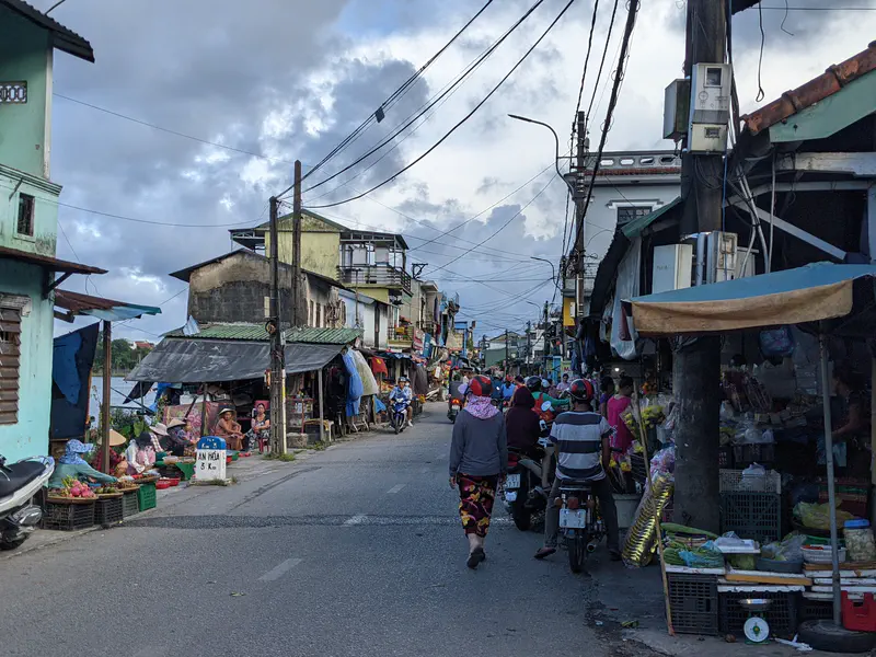 Bustling neighborhood street with shops, market stalls, and people walking or riding motorbikes.
