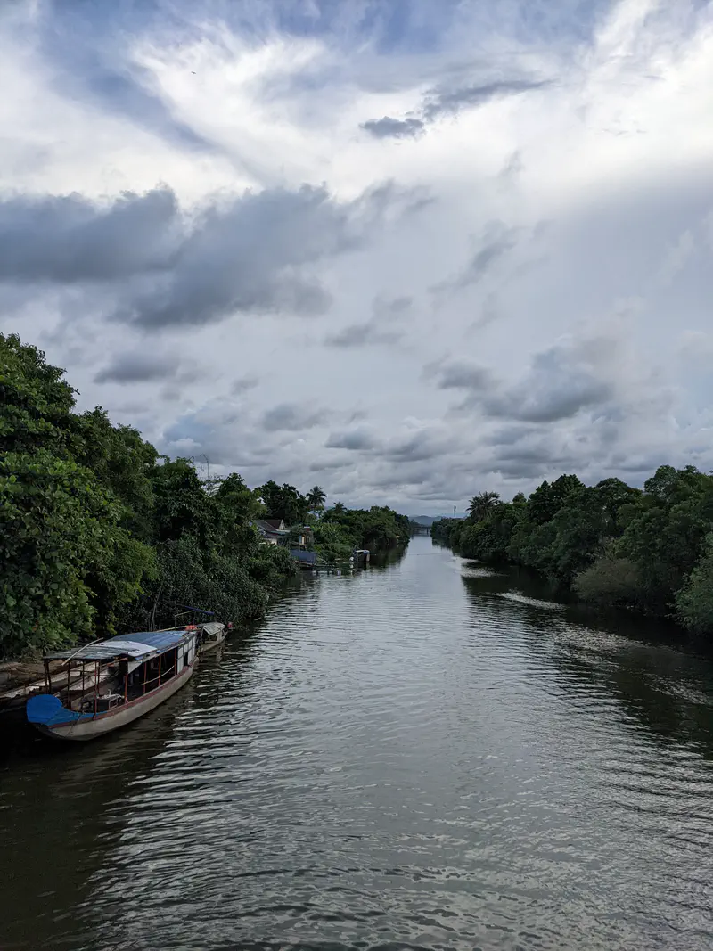 Calm river surrounded by lush green trees with boats docked along the bank under a cloudy sky.