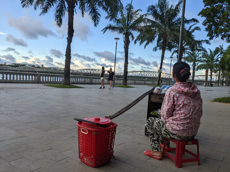 Woman sitting on a red stool by a riverside walkway with palm trees and a bridge in the background.