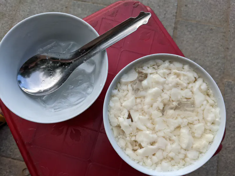 Two bowls on a red table, one with tofu pudding and ice, the other with ice cubes and a spoon.
