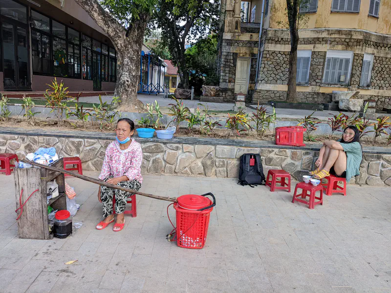 Street vendor sitting on a stool beside her setup with a red basket, while a woman sits nearby with food bowls.