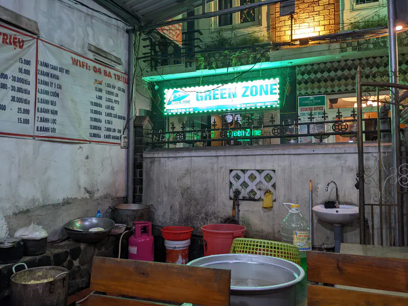 Outdoor food stall with cooking pots, gas canisters, and a menu on the wall beneath a glowing Green Zone sign.