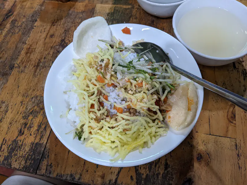Plate of rice topped with shredded vegetables, crispy noodles, and crackers beside a bowl of soup.