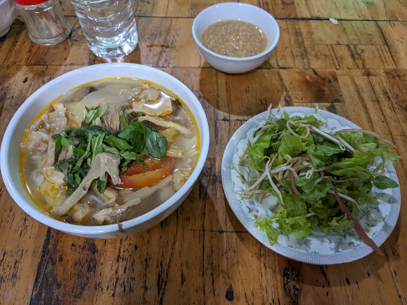 Bowl of noodle soup with vegetables and herbs served with a side plate of fresh greens and a small bowl of sauce.