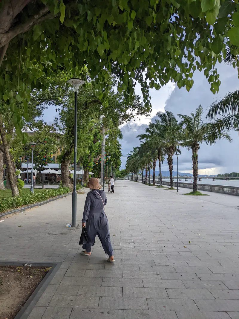 Person walking along a riverside promenade lined with trees and palm trees in Huế.