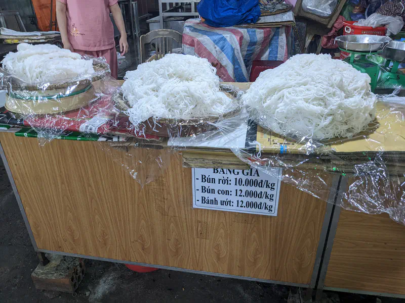 Bundles of fresh rice noodles displayed for sale at a market stall with prices listed.