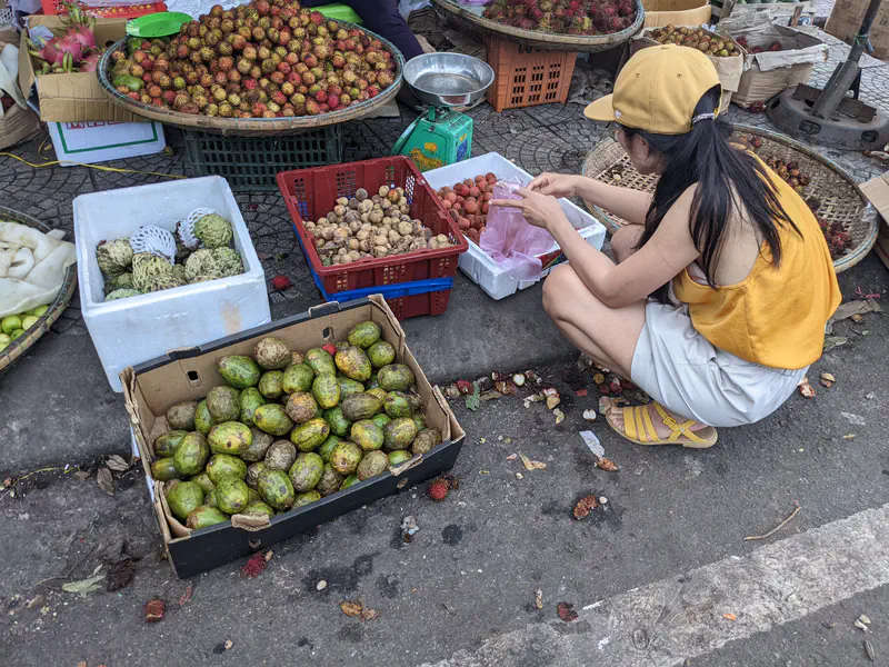 Woman in yellow top selecting tropical fruits at an outdoor market stall.