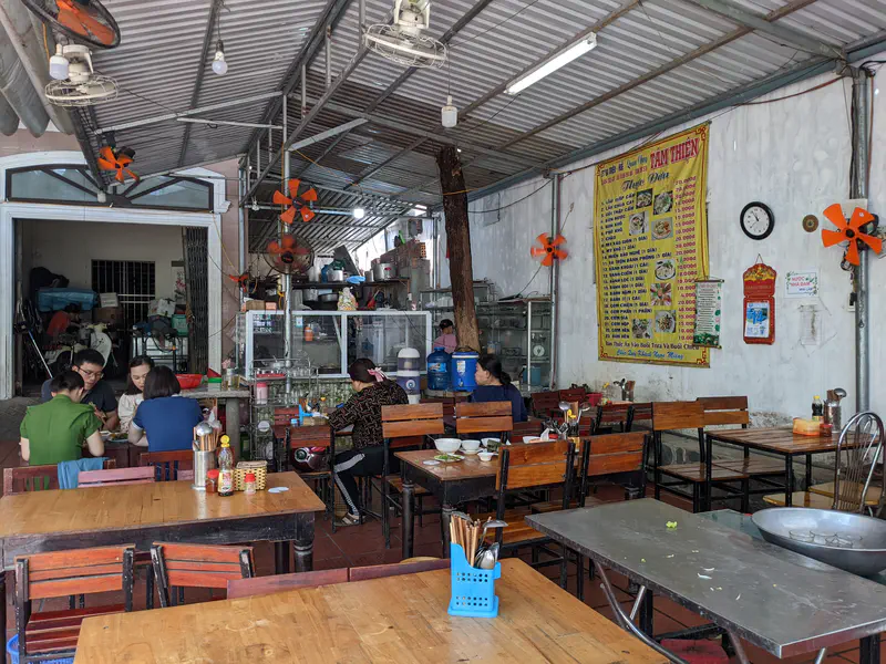 Casual Vietnamese eatery with wooden tables, wall menu, and people dining under ceiling fans.