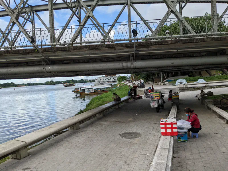Vendors and parked motorbikes under a bridge by a riverside walkway with boats on the water.