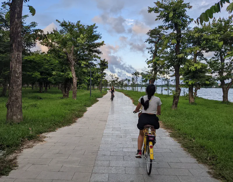Cyclists on a tree-lined path by the river in Huế during the evening.