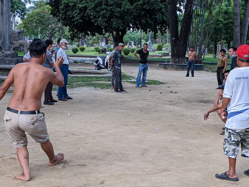 Group of men playing pétanque in a park, with one preparing to throw a ball.
