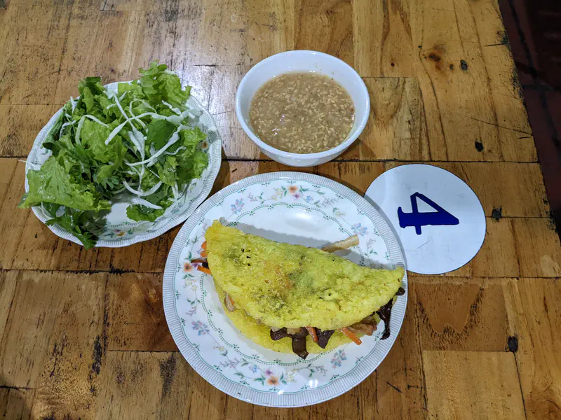 Plate with a Vietnamese crispy pancake filled with vegetables, served with lettuce, herbs, and dipping sauce.