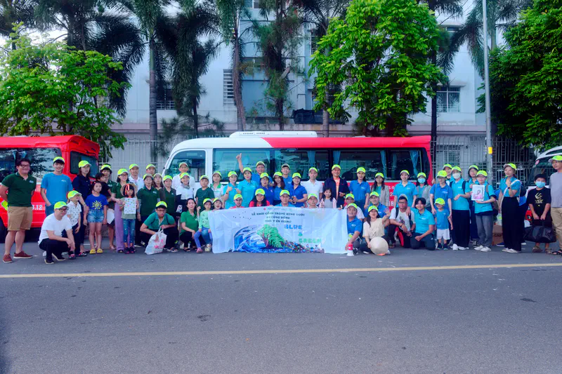 Large group of people in green caps posing with a Blue & Green banner in front of buses and trees.