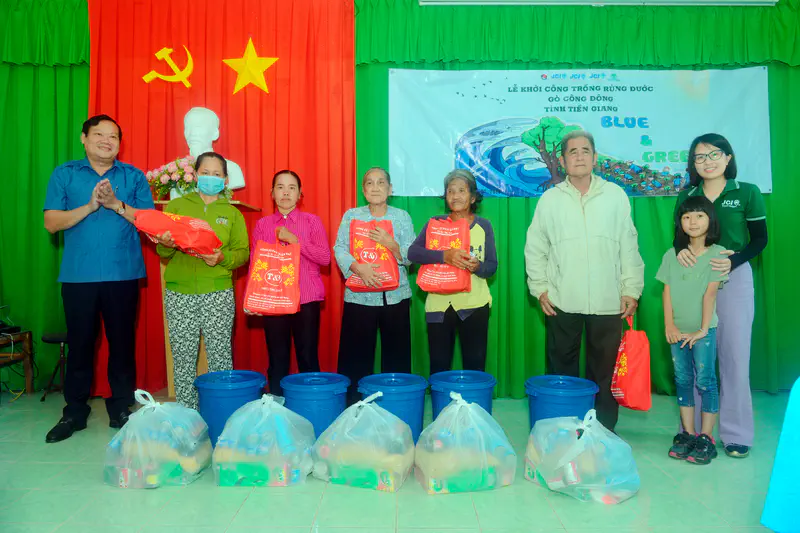 People standing on stage holding red gift bags with supplies in front of blue barrels and donation packages.