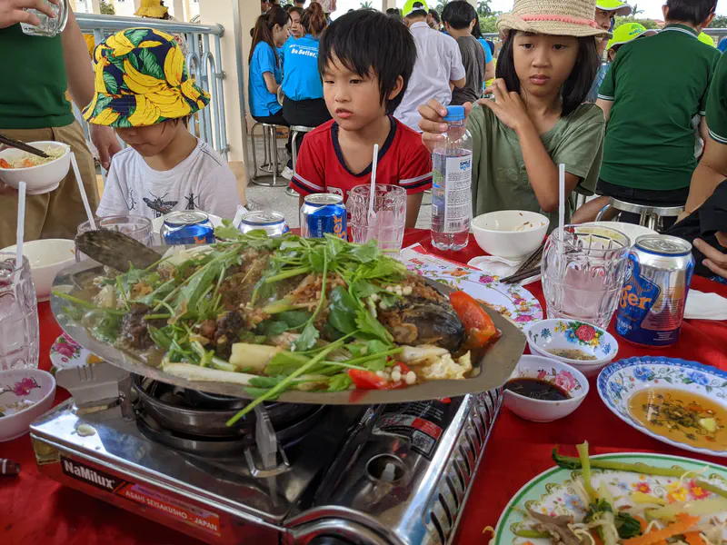 Children sitting around a table with a large fish dish cooked with vegetables and herbs on a portable gas stove, surrounded by bowls, drinks, and condiments.