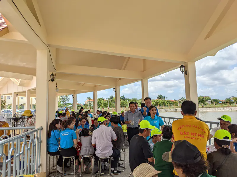 Large group of people eating together under a covered outdoor area by the water.