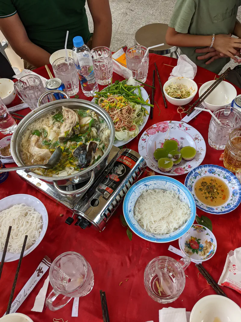 Table with hotpot, noodles, vegetables, and dipping sauces set for a shared meal.