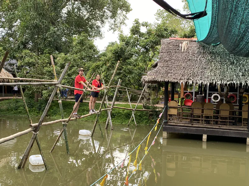 Two people in red shirts crossing a narrow bamboo bridge over a pond near a thatched-roof hut.