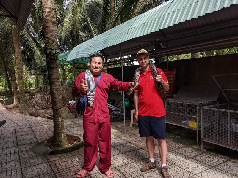 Two men smiling, one holding a python draped around his shoulders in an outdoor setting with palm trees.