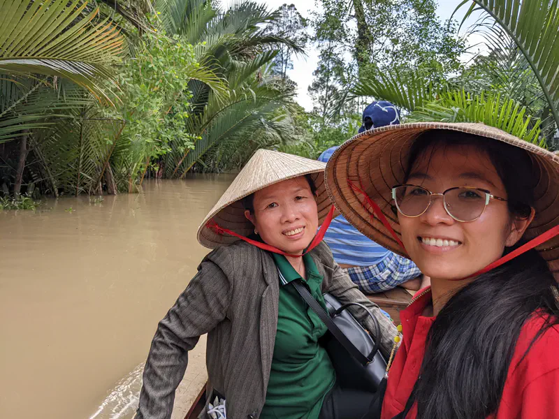 Two women wearing conical hats smiling on a boat ride through a canal lined with palm trees.