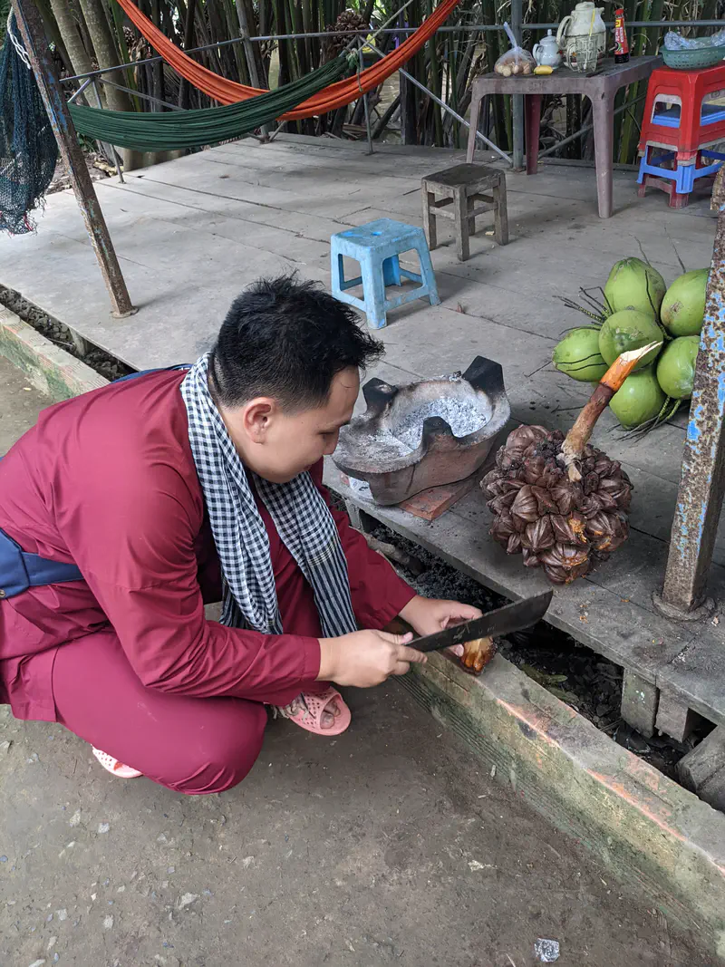 Man in traditional clothing using a knife to cut nipa palm fruit near a wooden platform.