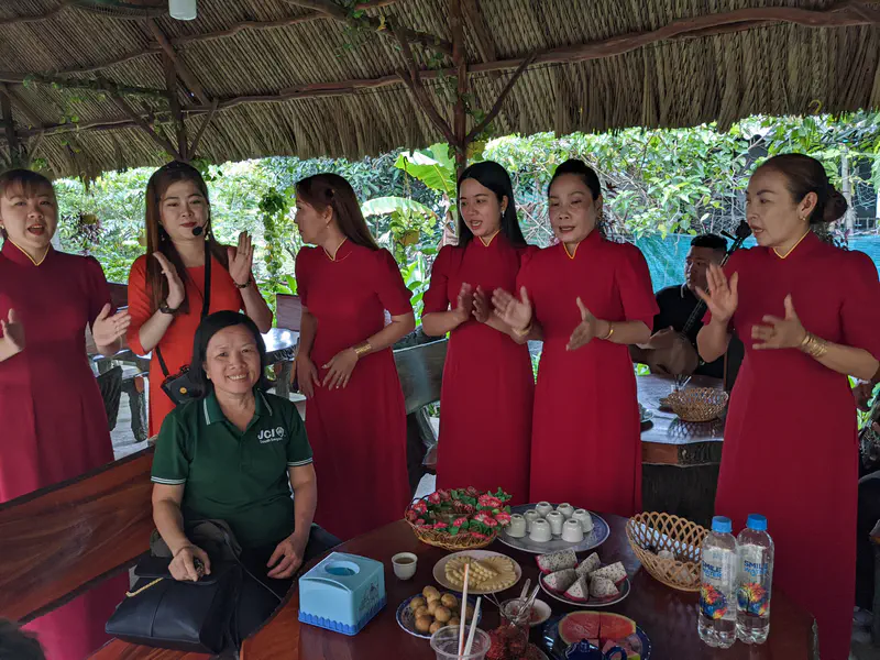 Group of women in red dresses singing beside a smiling woman seated at a table with fruit and tea.