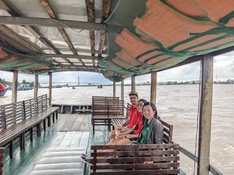 Three people sitting on wooden benches inside a covered boat on a wide river with a bridge in the distance.
