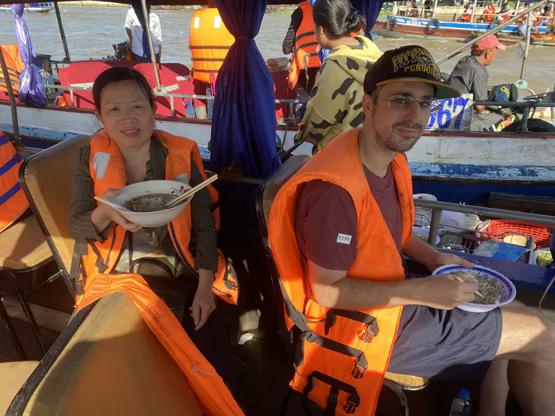 Two people wearing orange life jackets eating noodle soup on a boat at a floating market.