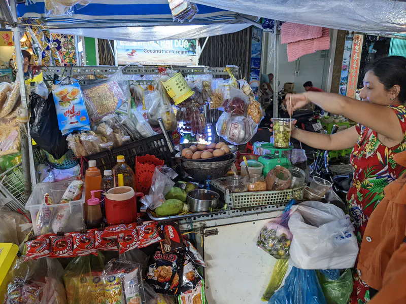 Street vendor preparing a snack at a small stall filled with eggs, sauces, and packaged goods.