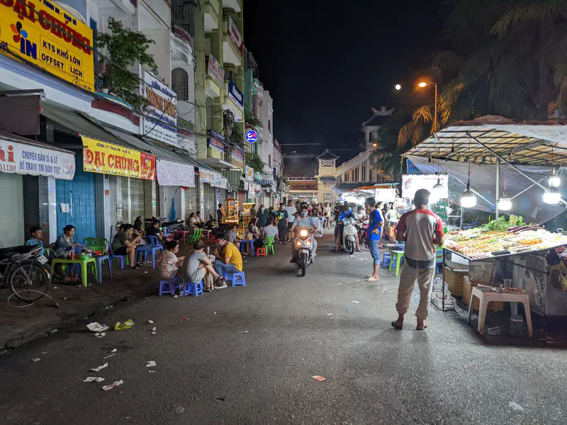 Bustling night market street with food stalls, diners on small stools, and motorbikes passing through.
