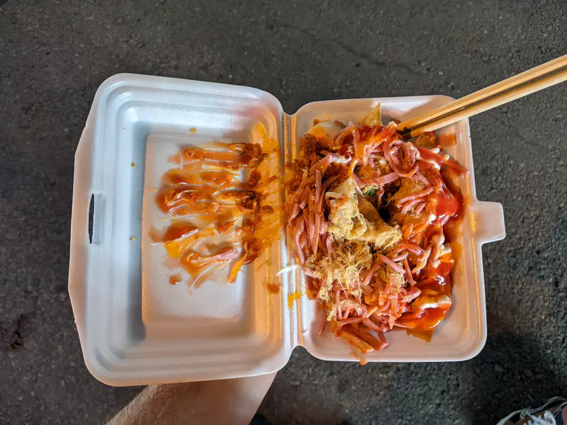 Takeaway box of Vietnamese street food topped with sausage, pork floss, mayonnaise, and chili sauce.